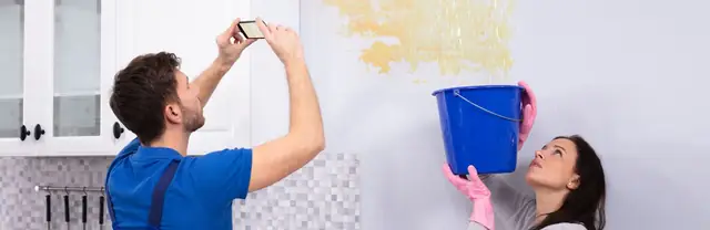 Homeowners photographing a water-stained wall while a bucket catches a leak